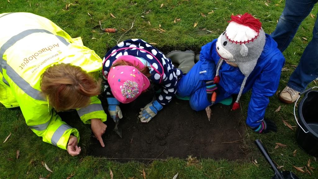 Two children and an archaeologist around a 1m by 1m test pit excavation