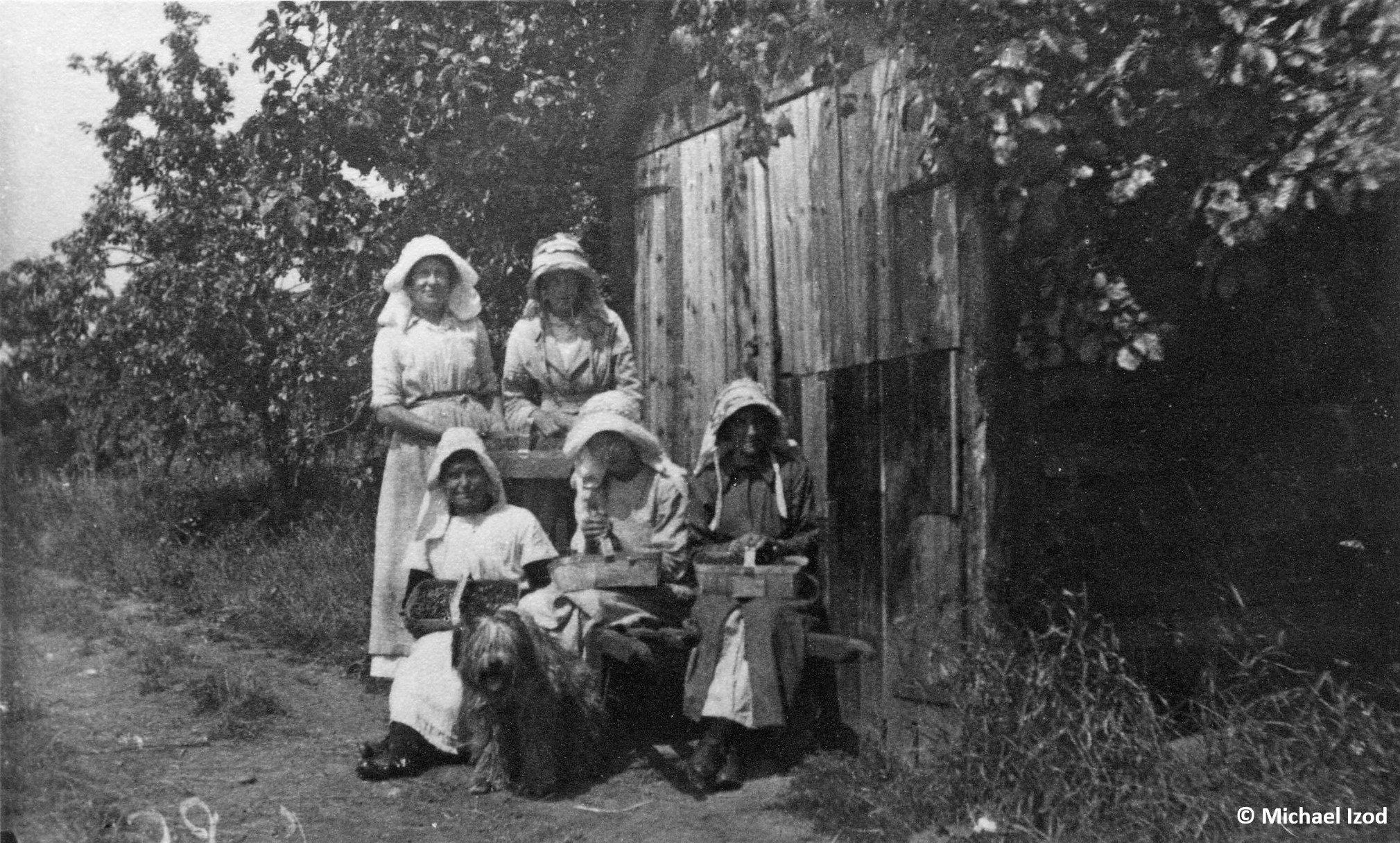 Five women with baskets of fruit by a wooden shed in an orchard