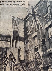 Image of photograph from Worcester Newspaper showing flags hanging from the upper floors of shops. A man is hanging precariously from a ledge to fix them.