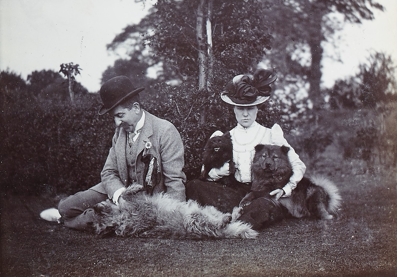 Black and white photograph of a couple sat outside on the grass with their dogs