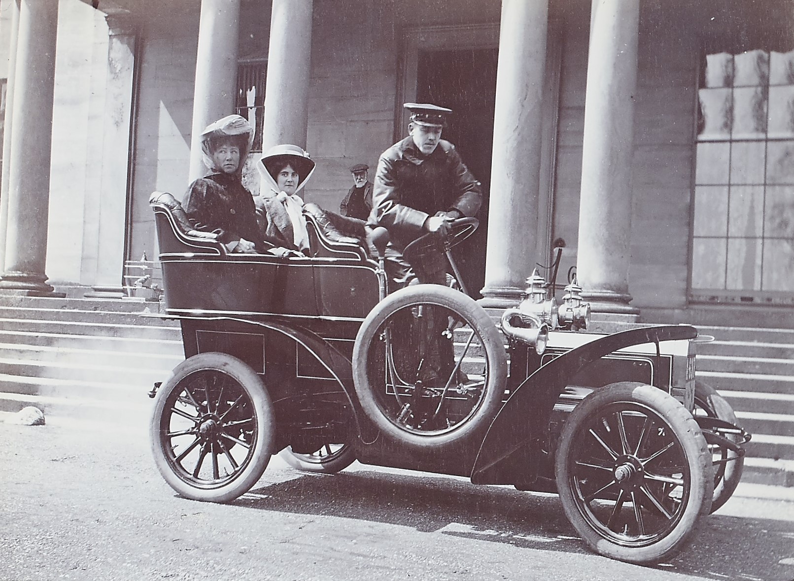 Black and white photograph of three people in hats sat in an old fashioned car outside