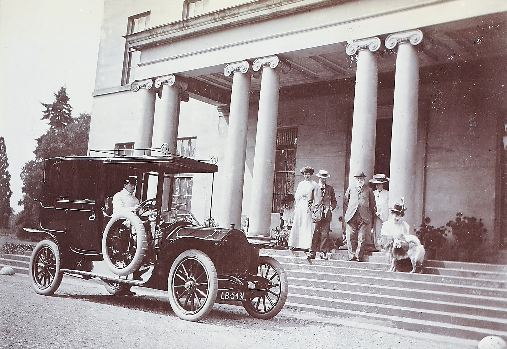 Black and white photograph of people on the steps of a grand country house with old fashioned car in front