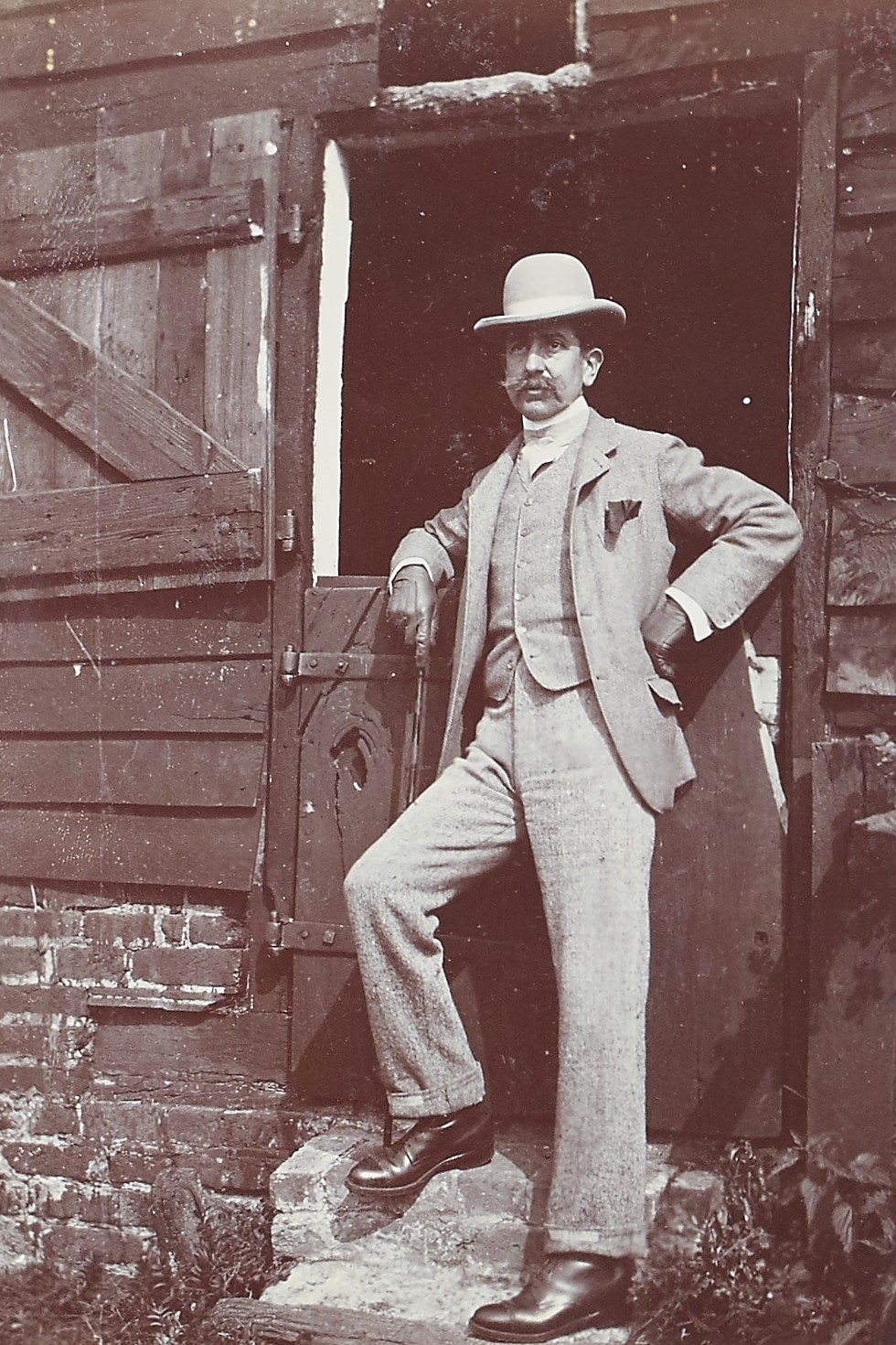 Black and white photograph of a man stood by a stable door wearing a suit and bowler hat