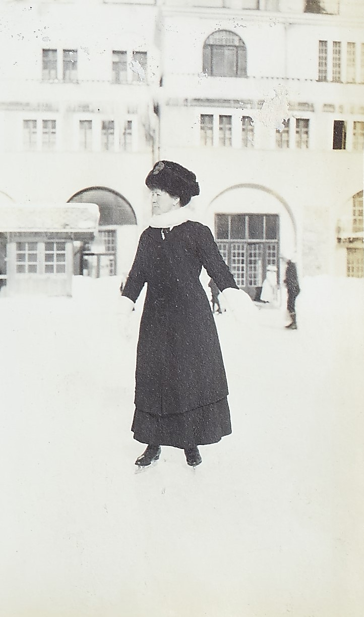 Black and white photograph of a women in a coat and fur hat standing on an ice rink