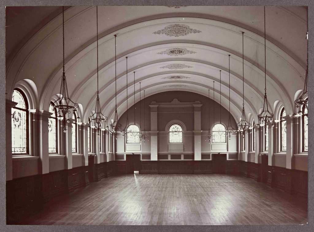Black and white photograph of the ballroom at Barnsley Hall which features an oval plastered ceiling with decorations, iron light fittings and many large windows running the length of the room, along with a smooth laminate wooden floor
