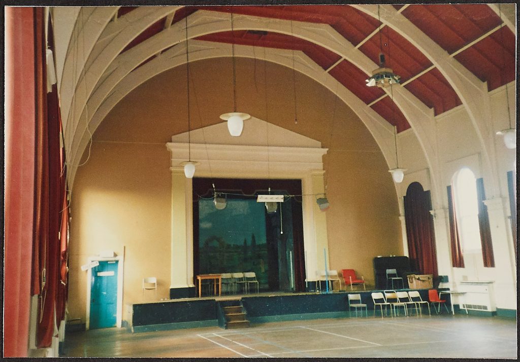 Coloured photograph showing the interior of the Ballroom at Powick featuring a stage with a wooden ceiling and smooth floor dated 1989 prior to Powick Hospital closing